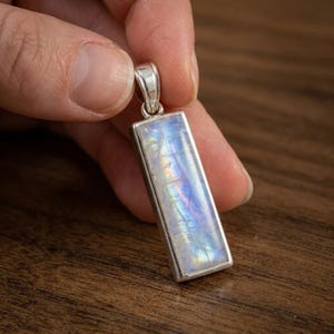 May include: A rectangular pendant with a shimmering, iridescent moonstone set in a silver-colored frame. The moonstone displays a play of colors, including blues, greens, and yellows. The pendant is held against a dark wooden surface.