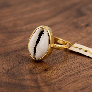 Puede incluir: Un anillo de tono dorado que presenta una concha de cauri blanca central con una abertura dentada de color marrón oscuro. La banda es lisa y metálica, y una pequeña etiqueta de papel rectangular está unida al lado con un hilo dorado.