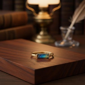 May include: A gold ring featuring a rectangular gemstone with a shimmering, multi-coloured effect. The gemstone displays blues, greens, and yellows. The ring sits on a dark wooden surface, with a blurred background of books and a lamp.