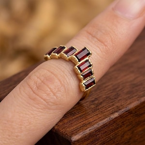Pode incluir: Um anel dourado com uma série de pedras preciosas retangulares de cor vermelha escura. O design do anel é uma fileira curva de pedras, cravadas numa banda dourada texturizada. As pedras são espaçadas uniformemente.