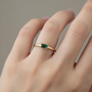 May include: A gold ring featuring a rectangular, emerald-cut green gemstone. The ring has a thin, polished gold band. The gemstone is set horizontally, creating a minimalist and elegant design.