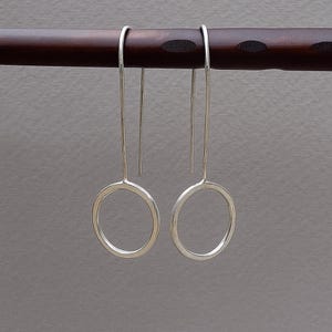 Op de afbeelding: Een paar zilverkleurige, minimalistische oorbellen. Elke oorbel heeft een lange, dunne draad die verbonden is met een cirkelvormige ring. De oorbellen worden gepresenteerd tegen een donkerbruine houten staaf en een grijze achtergrond.