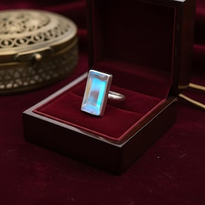 May include: A silver ring featuring a rectangular, iridescent gemstone, presented in a dark wooden jewellery box with a red velvet lining. The gemstone reflects blue and white colours. An ornate, gold-coloured jewellery box is in the background.