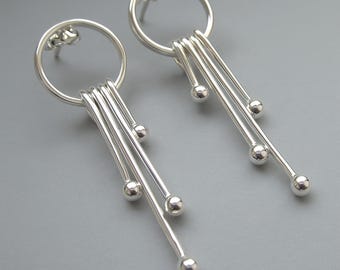 Peut inclure: Une paire de boucles d'oreilles pendantes argentées. Chaque boucle d'oreille est composée d'un cercle relié à des barres fines et droites avec de petites perles rondes. Design moderne et minimaliste.