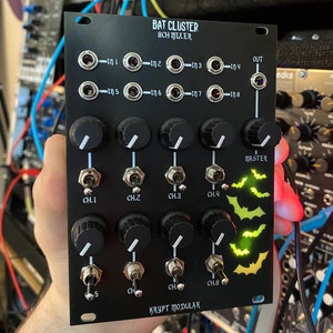 May include: A black 8-channel mixer with the text "BAT CLUSTER 8CH MIXER" and "KRYPT MODULAR" printed on the front. The mixer has eight knobs, eight switches, and eight input jacks. The mixer has a master volume knob and a master switch. The mixer has a green LED that is lit up in the shape of a bat.
