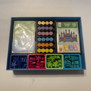 May include: A board game organiser containing game pieces. It features cards, multi-coloured hexagonal tiles, and flower-shaped tokens in blue, red, green, and teal. The game box also has a card with the text "Mangini Estate".