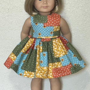 May include: A doll with blonde hair and blue eyes wearing a sleeveless dress featuring a patchwork pattern of green, blue, orange, and yellow fabric squares with floral and checkered prints, including small sunflower and white daisy motifs.