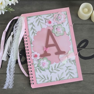 May include: A pink scrapbook with a spiral binding, decorated with floral embellishments and the letter "A". The cover features a scalloped pink shape, floral accents, and a bow. Ribbons and lace hang from the binding.