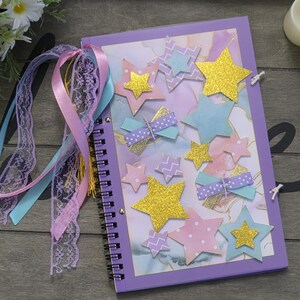 May include: A spiral-bound journal with a lavender cover adorned with star-shaped embellishments in gold glitter, pastel blue, and pink. The journal is tied with ribbons and lace in shades of blue, pink, and lavender. A decorative item for writing or sketching.