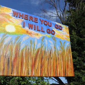 Może przedstawiać: Ręka trzyma namalowane płótno z polem wysokiej trawy i błękitnym niebem. Na płótnie widnieje napis "Where you go I will go Ruth 1:16" czerwonymi i niebieskimi literami.