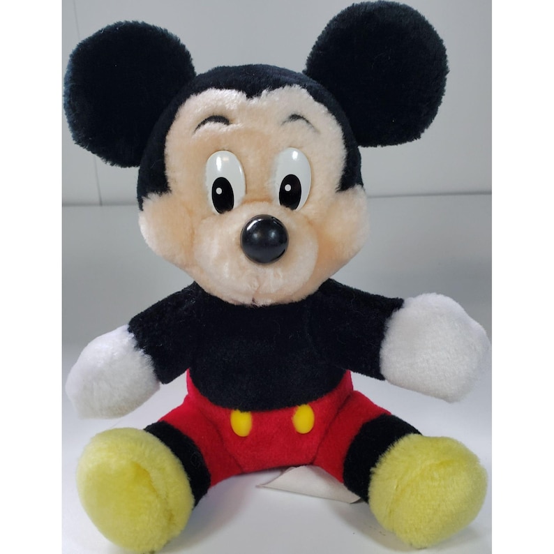 May include: A plush Mickey Mouse toy with black ears, a black body, red shorts with yellow buttons, and yellow shoes. The toy has a friendly expression with large eyes and a black nose.