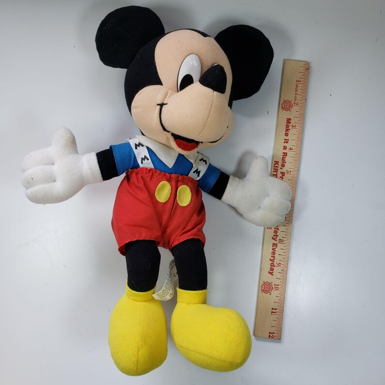 May include: A plush Mickey Mouse toy wearing a blue and white striped shirt with a red and white overall. The toy is approximately 30 cm tall and has a yellow nose and black eyes.