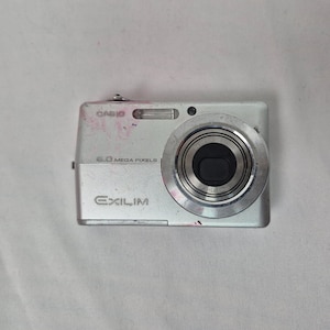 May include: Silver Casio Exilim digital camera with a lens, flash, and the text "6.0 MEGA PIXELS". The camera has some pink marks on the top and is on a white surface.