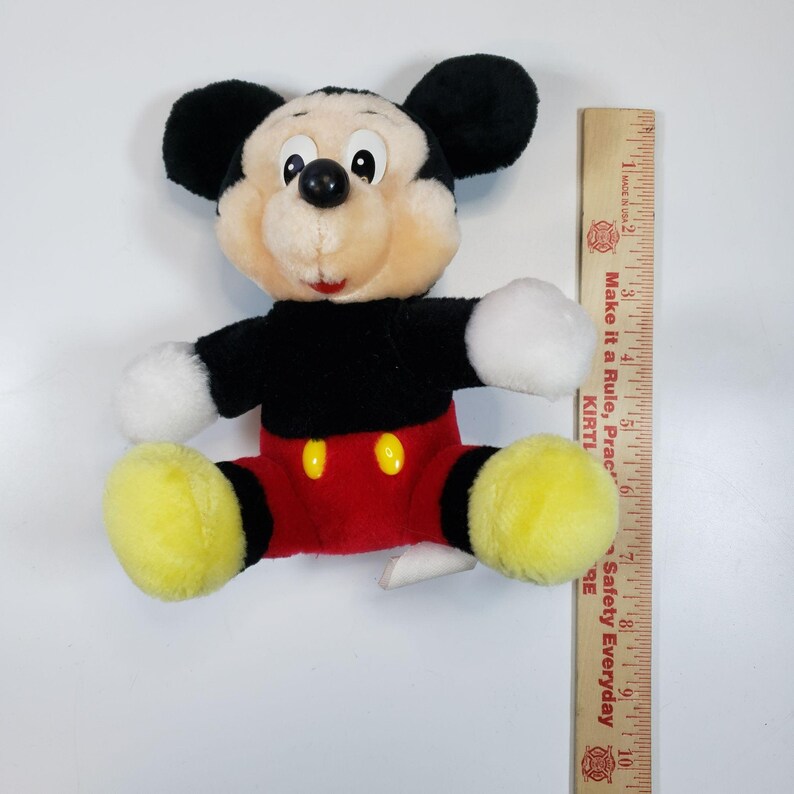 May include: A plush Mickey Mouse toy sitting with his legs crossed. He is wearing red shorts with black trim and yellow shoes. He has a black nose and big, round eyes.