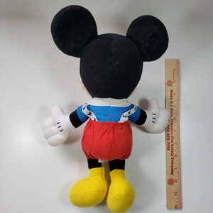 May include: A plush Mickey Mouse toy wearing red overalls with blue suspenders and a blue and white striped shirt. The toy is standing on a white surface with a ruler in the background.