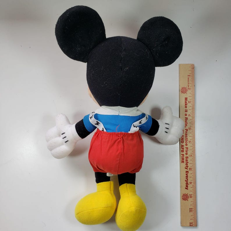 May include: A plush Mickey Mouse toy wearing red overalls with blue suspenders and a blue and white striped shirt. The toy is standing on a white surface with a ruler in the background.
