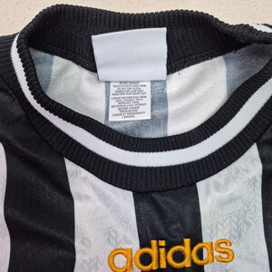 Puede incluir: Primer plano de una camiseta Adidas a rayas blancas y negras. La camiseta tiene un cuello negro con rayas blancas y el logo de Adidas en dorado. Se ve una etiqueta blanca con instrucciones de cuidado.