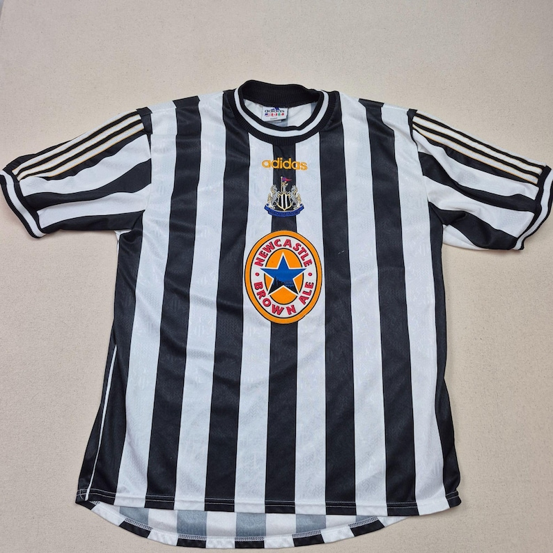 Puede incluir: Camiseta de f&uacute;tbol a rayas blancas y negras con el logo de Newcastle Brown Ale. La camiseta tiene cuello redondo y mangas cortas con tres rayas en los hombros. El logo de Adidas es visible.