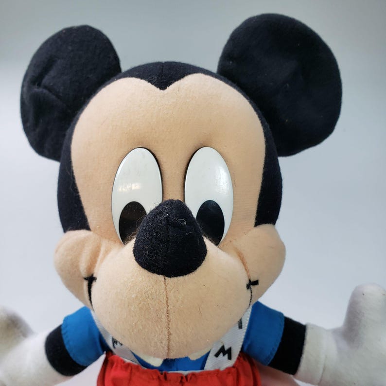 May include: A plush Mickey Mouse toy wearing a blue shirt and red dungarees. The toy has large black ears, white eyes, and a black nose.