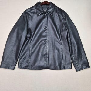 May include: A black leather jacket with a collared neckline and button-down closure. The jacket has long sleeves and a tailored fit. The jacket is hanging on a wooden hanger against a neutral background.