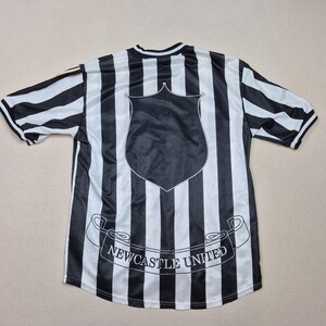 Puede incluir: Camiseta de f&uacute;tbol a rayas blancas y negras con un escudo y la inscripci&oacute;n "Newcastle United" en una pancarta. La camiseta tiene mangas cortas y cuello redondo. Las rayas son verticales.