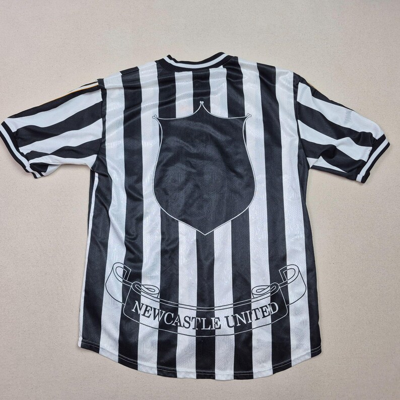 Puede incluir: Camiseta de f&uacute;tbol a rayas blancas y negras con un escudo y la inscripci&oacute;n "Newcastle United" en una pancarta. La camiseta tiene mangas cortas y cuello redondo. Las rayas son verticales.