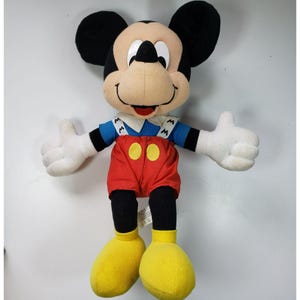 May include: A plush Mickey Mouse toy wearing a blue shirt with white braces and red shorts. The toy has black shoes and a big smile.