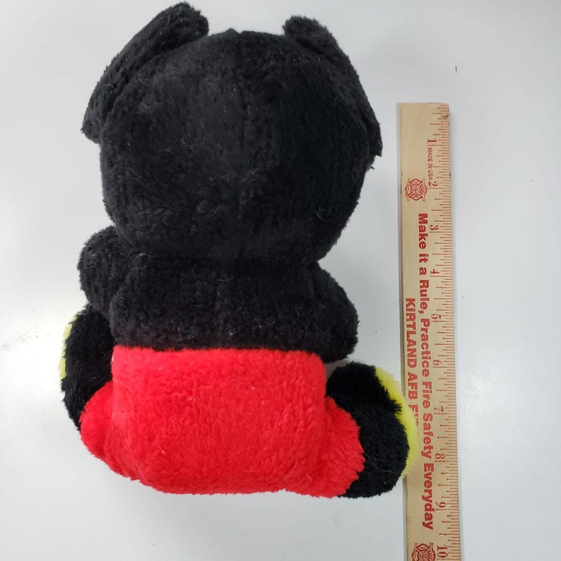 Pode incluir: Brinquedo de pel&uacute;cia preto e vermelho com detalhes amarelos. O brinquedo tem a forma de um personagem de desenho animado com orelhas grandes e corpo redondo. O brinquedo est&aacute; sentado em uma superf&iacute;cie branca.
