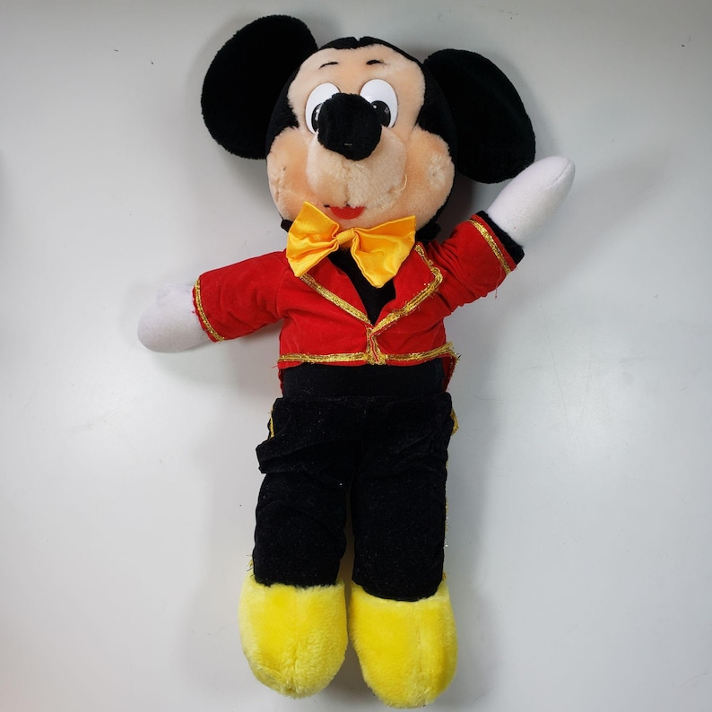 May include: A plush Mickey Mouse toy wearing a red and gold jacket with a yellow bow tie. The toy has black ears, black shoes, and yellow feet.