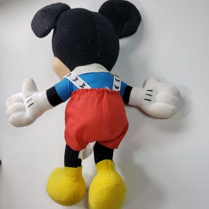 May include: A plush Mickey Mouse toy wearing a blue shirt with white braces and red trousers. The toy has black ears, white gloves, and yellow shoes.