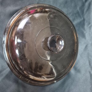 May include: A round, transparent, smoke-tinted glass lid for cookware. The lid features a central, raised circular knob handle and a stepped rim design. The word Pyrex is embossed along the outer edge of the glass surface.