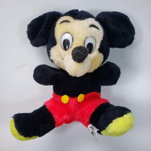 May include: A vintage plush Mickey Mouse toy with black fur, large eyes, and a red and yellow outfit. The toy is sitting on a white surface.