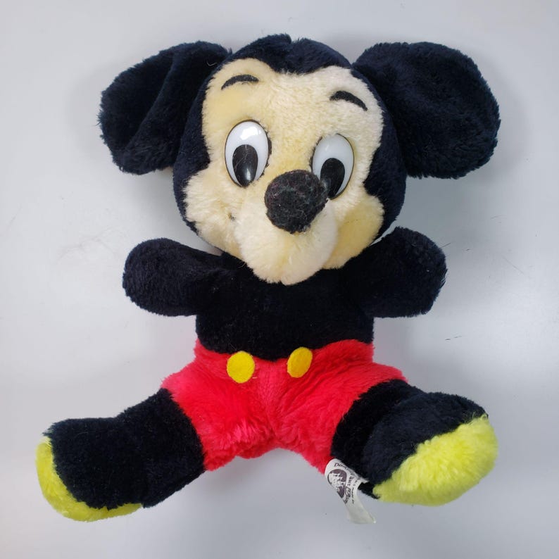 May include: A vintage plush Mickey Mouse toy with black fur, large eyes, and a red and yellow outfit. The toy is sitting on a white surface.
