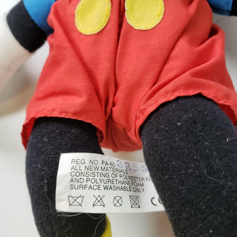 May include: A red and black plush toy of Mickey Mouse. The toy is wearing red shorts with yellow buttons and has a black and white face. The toy has a label on the bottom that reads "REG. NO. PA-60 3355 ALL NEW MATERIALS CONSISTING OF POLYESTER FI AND POLYURETHANE FOAM SURFACE WASHABLE ONLY".