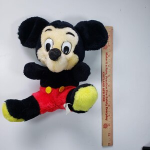 May include: A black and yellow plush Mickey Mouse toy wearing red shorts with a yellow button. The toy is sitting on a white surface with a ruler in the background.