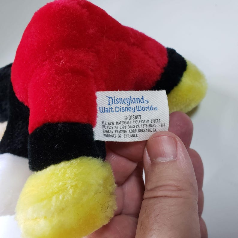 May include: A red, black, and yellow plush Mickey Mouse toy with a Disneyland Walt Disney World tag. The tag reads "Disneyland Walt Disney World" and "DISNEY ALL NEW MATERIALS POLYESTER FIBERS ME 1575 PA 1378 OHIO PA 1378 MASS T-616 CARASA TRADING CORP. BURBANK, CA PRODUCT OF SRI LANKA."
