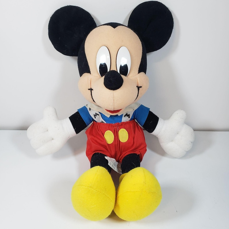 May include: A plush Mickey Mouse toy wearing a blue and white shirt, red overalls with yellow buttons, and yellow shoes. The toy has black ears, a black nose, and large white eyes.