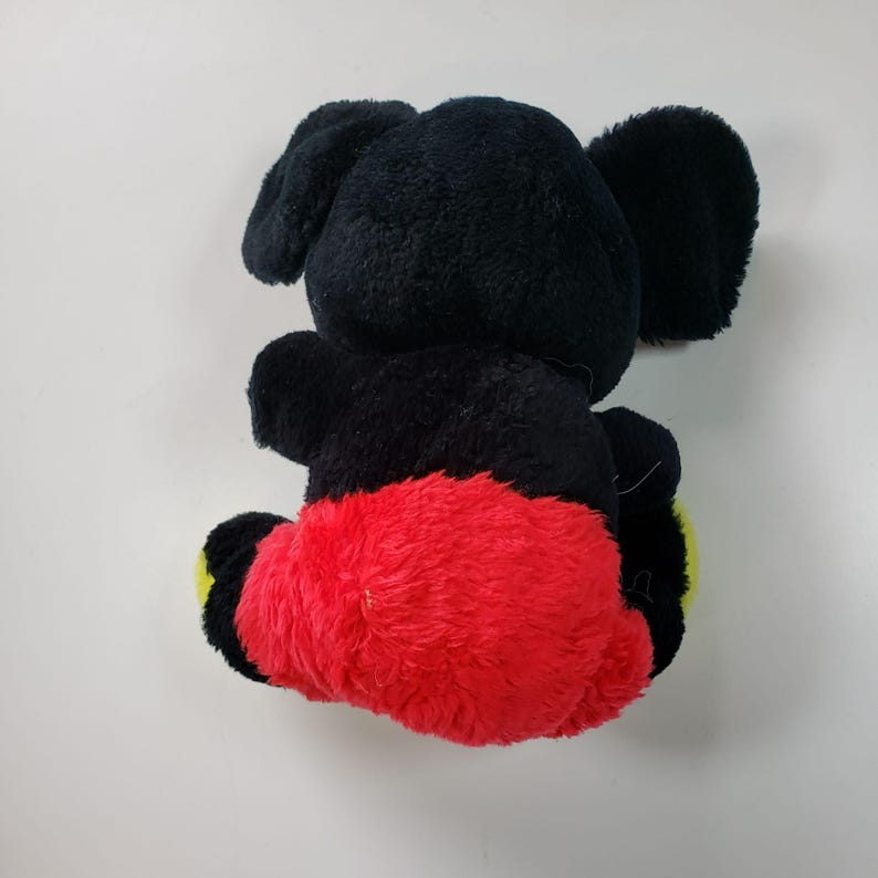May include: Black and red plush toy with large ears and yellow accents. The toy is sitting on a white surface.