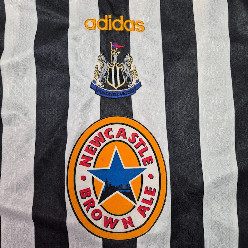 Puede incluir: Primer plano de una camiseta de f&uacute;tbol a rayas blancas y negras. El logotipo de Adidas est&aacute; bordado en dorado. El escudo del Newcastle United est&aacute; encima de un logotipo circular que dice "Newcastle Brown Ale".