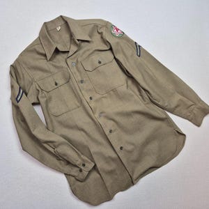 May include: A long-sleeved tan button-down shirt featuring two chest pockets, a pointed collar, and buttoned cuffs. The left shoulder displays a circular patch with a red star and the letter A, while both sleeves feature dark blue chevron stripes.