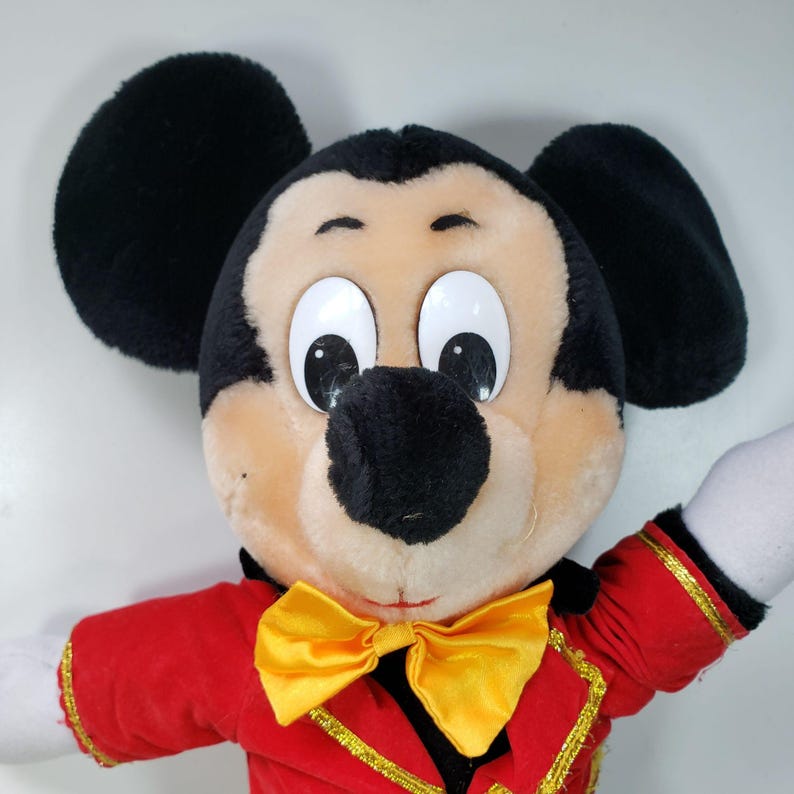 May include: A plush Mickey Mouse toy wearing a red jacket with gold trim and a yellow bow tie. The toy has black ears, a black nose, and large white eyes.