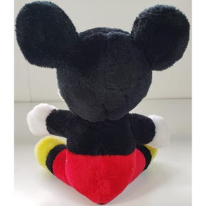 May include: A black and red plush Mickey Mouse toy sitting on a white surface. The toy has large black ears, a black body, red shorts, and yellow shoes.