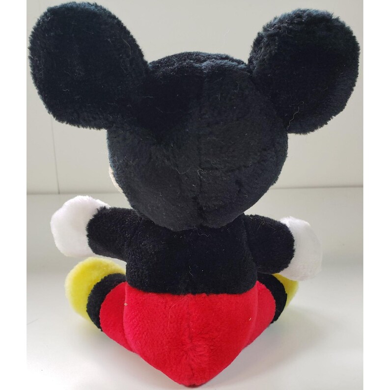 May include: A black and red plush Mickey Mouse toy sitting on a white surface. The toy has large black ears, a black body, red shorts, and yellow shoes.