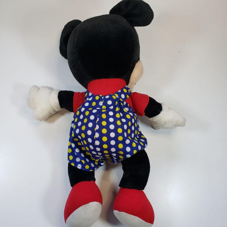 K&ouml;nnte beinhalten: Ein Pl&uuml;sch-Mickey-Mouse-Spielzeug, das ein rotes Hemd und eine blaue Latzhose mit wei&szlig;en und gelben Punkten tr&auml;gt. Das Spielzeug hat schwarzes Fell und gro&szlig;e Ohren.