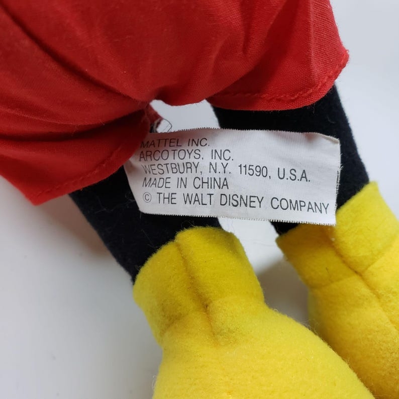 May include: A plush Mickey Mouse toy with red shorts, black shoes, and yellow gloves. The toy has a tag that reads "MATTEL INC. ARCO TOYS, INC. WESTBURY, N.Y. 11590, U.S.A. MADE IN CHINA &copy; THE WALT DISNEY COMPANY"