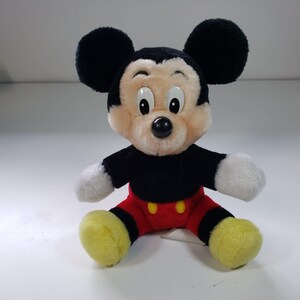 May include: A plush Mickey Mouse toy with black ears, a black body, red shorts with yellow buttons, and yellow shoes. The toy has a black nose and white eyes.