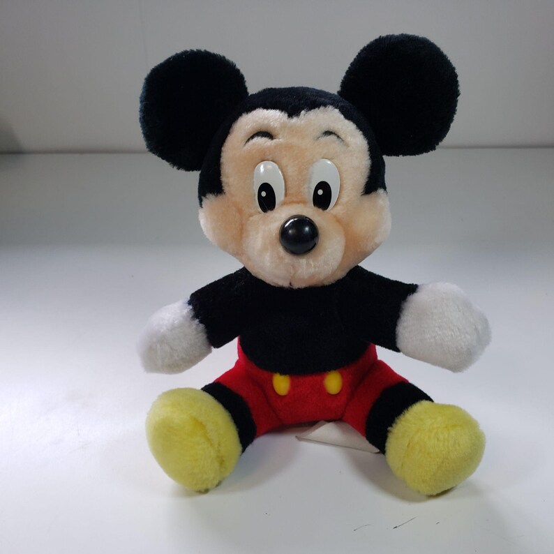 May include: A plush Mickey Mouse toy with black ears, a black body, red shorts with yellow buttons, and yellow shoes. The toy has a black nose and white eyes.