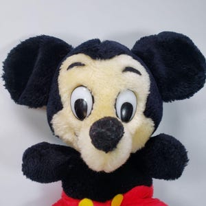 May include: A plush Mickey Mouse toy with black ears, a black nose, and large white eyes. The toy is wearing red shorts with yellow buttons.