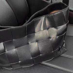 Puede incluir: Un bolso de mano de cuero negro con un dise&ntilde;o tejido. El bolso es rectangular, con una abertura ancha y un aspecto suave y texturizado. El bolso est&aacute; sobre un asiento de cuero negro.