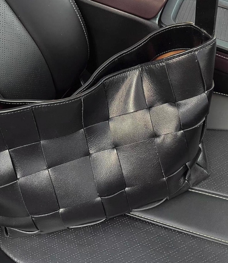 Puede incluir: Un bolso de mano de cuero negro con un dise&ntilde;o tejido. El bolso es rectangular, con una abertura ancha y un aspecto suave y texturizado. El bolso est&aacute; sobre un asiento de cuero negro.